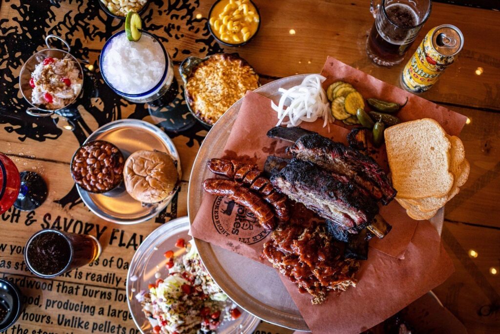 A table filled with barbecue foods, including ribs, sausage, pickles, onions, white bread, baked beans, macaroni and cheese, corn, a sandwich, drinks, and various sides, viewed from above.