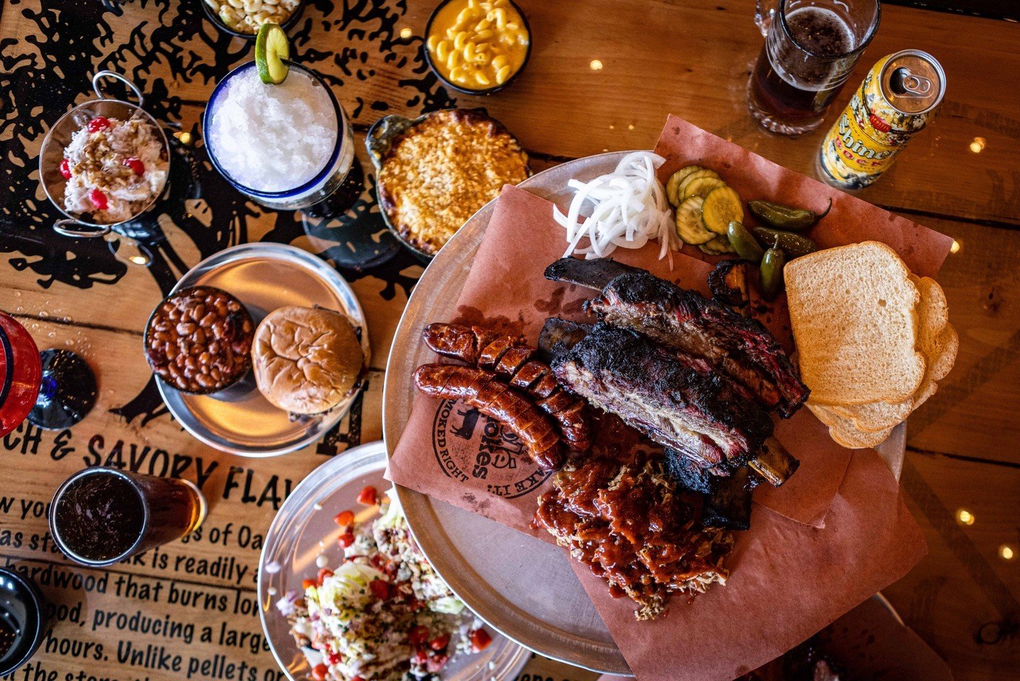 A table filled with barbecue foods, including ribs, sausage, pickles, onions, white bread, baked beans, macaroni and cheese, corn, a sandwich, drinks, and various sides, viewed from above.