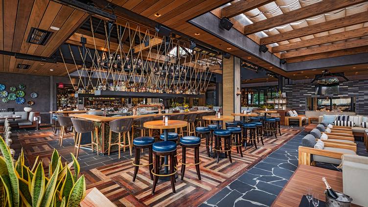Modern restaurant interior with wooden ceiling beams, patterned wooden flooring, and stylish hanging lights. The space features a central bar with blue barstools, high-top tables, cushioned seats, and large windows for natural light.