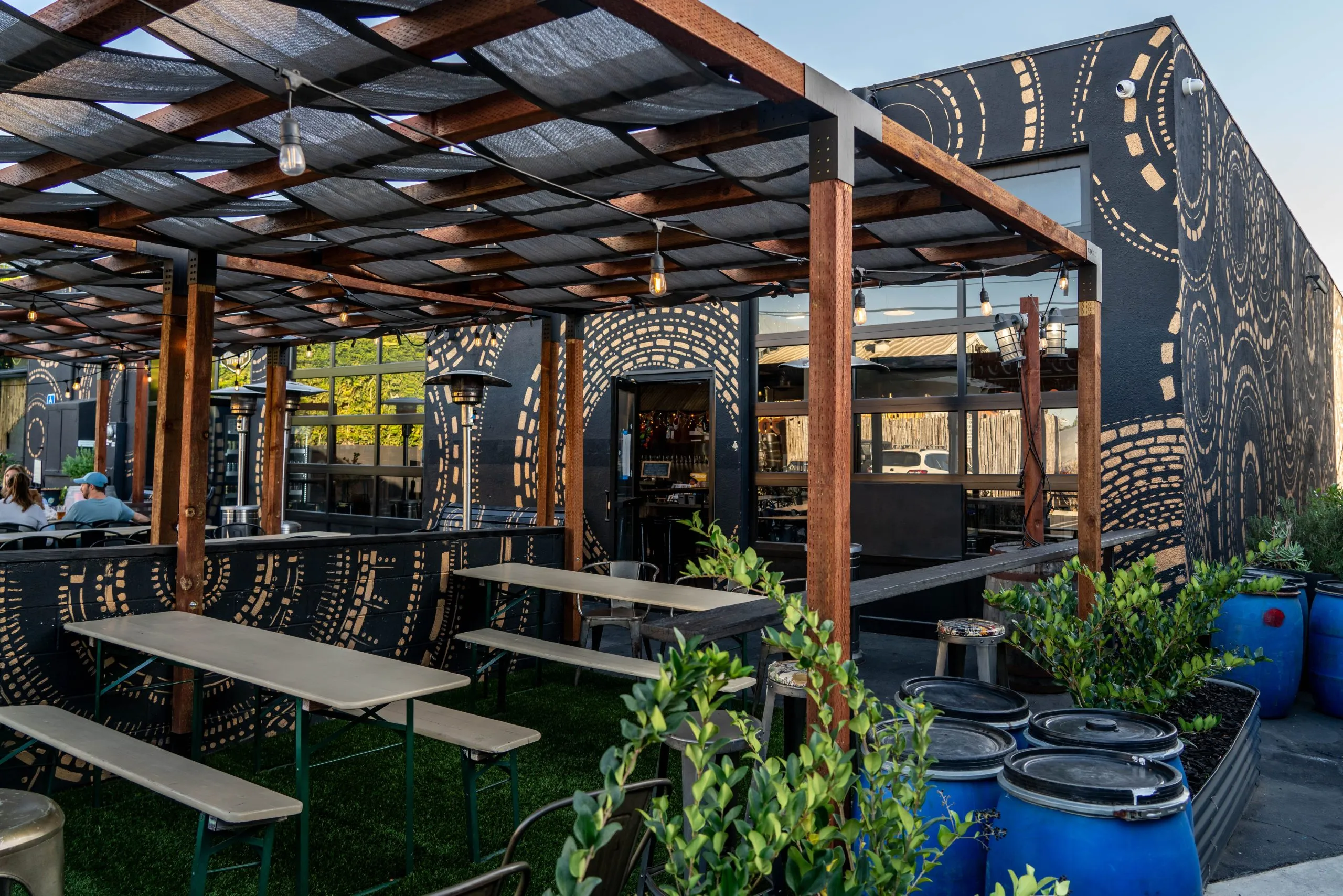 Outdoor seating area with wooden tables and benches under a pergola, next to a building with artistic, circular patterns on black walls. Blue barrels with plants line the side, and string lights hang above.