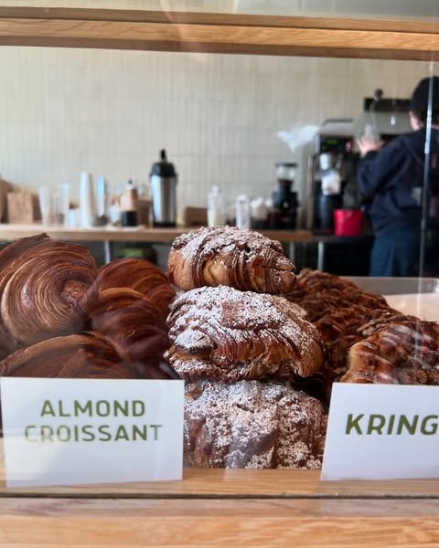 Almond croissants topped with powdered sugar are stacked behind a glass display at a bakery, with a sign reading ALMOND CROISSANT in front and a blurred barista working in the background.