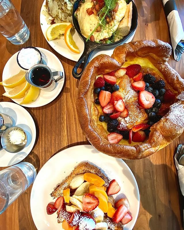 A wooden table set with a Dutch baby pancake topped with fresh berries, toast with fruit, a skillet omelet with potatoes, lemon wedges, syrup, butter, and glasses of water.