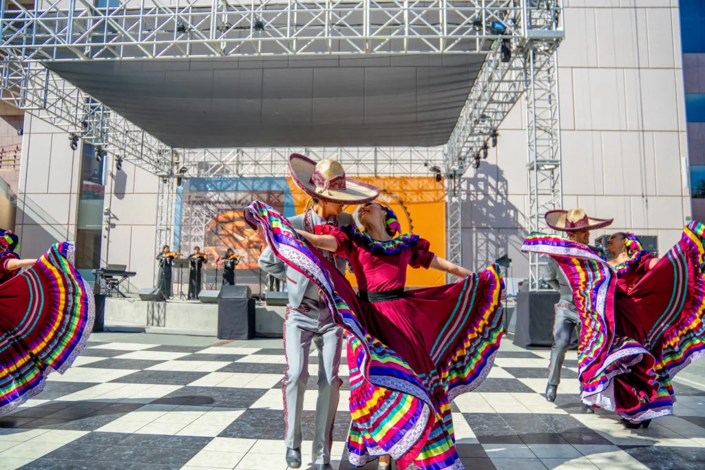 Dancers in vibrant costumes perform traditional Mexican folklórico on a checkered outdoor stage, with musicians playing in the background and a metal scaffold structure overhead.
