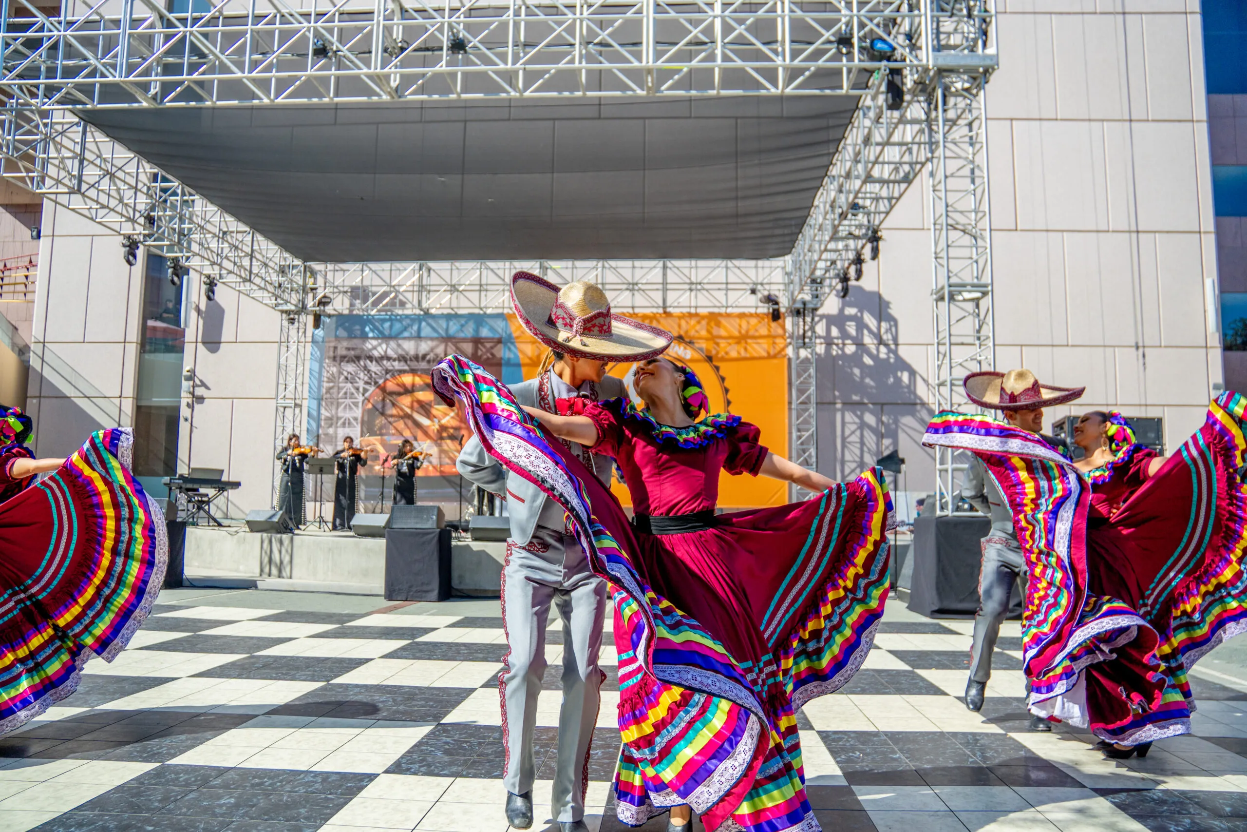 Dancers in vibrant costumes perform traditional Mexican folklórico on a checkered outdoor stage, with musicians playing in the background and a metal scaffold structure overhead.