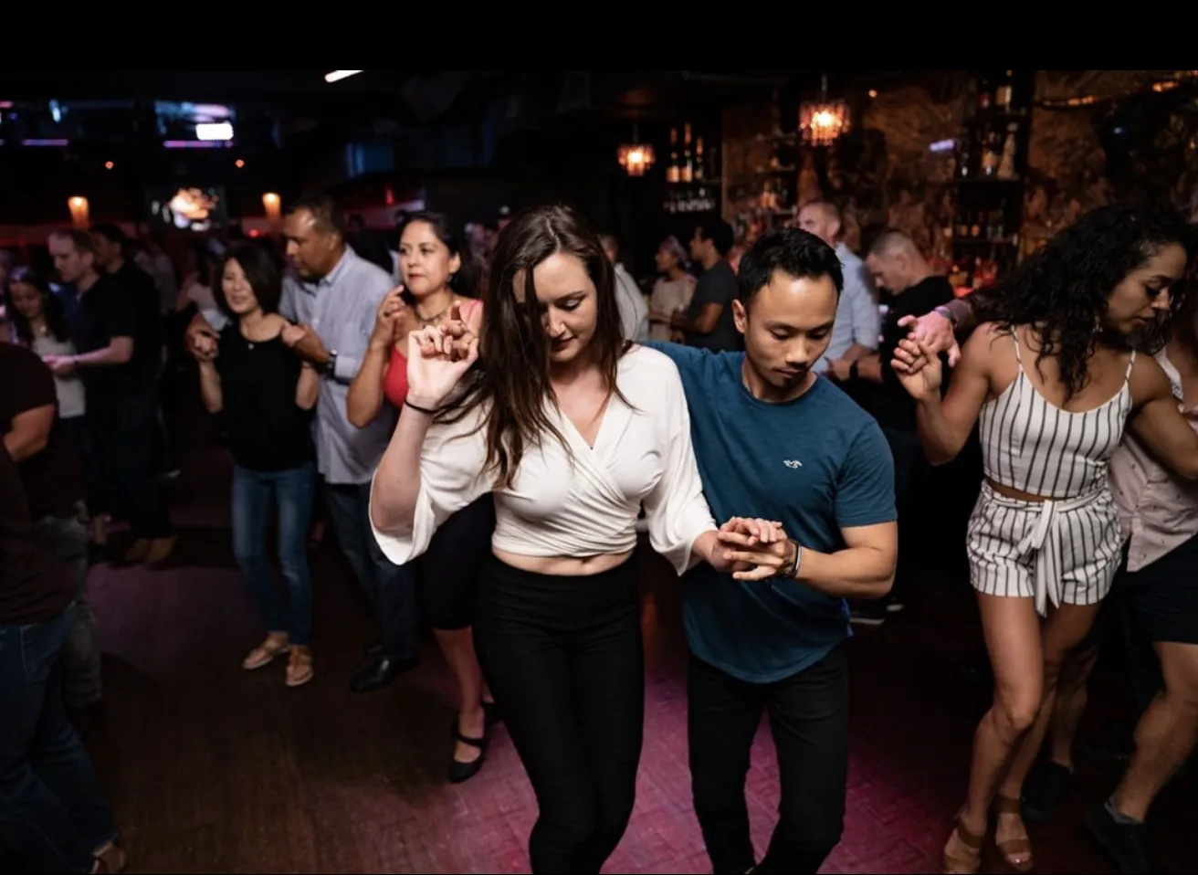 A group of people dance closely in pairs in a dimly lit bar or club, focusing on their movements. The atmosphere is lively and social, with couples holding hands and enjoying the music together.