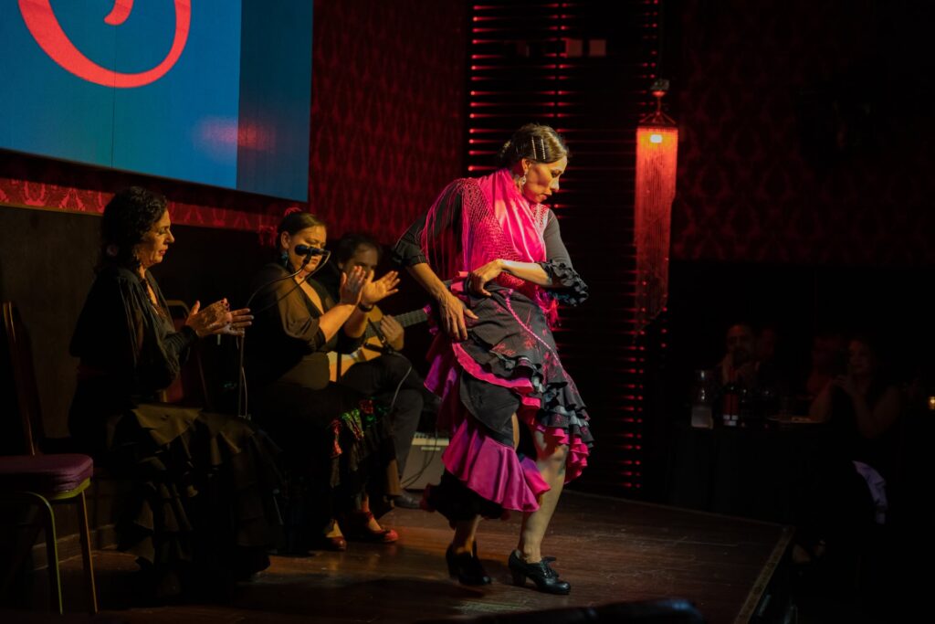 A flamenco dancer in a black and pink dress performs on stage, while seated musicians and singers in dark outfits clap along. The audience watches from tables in a dimly lit venue.