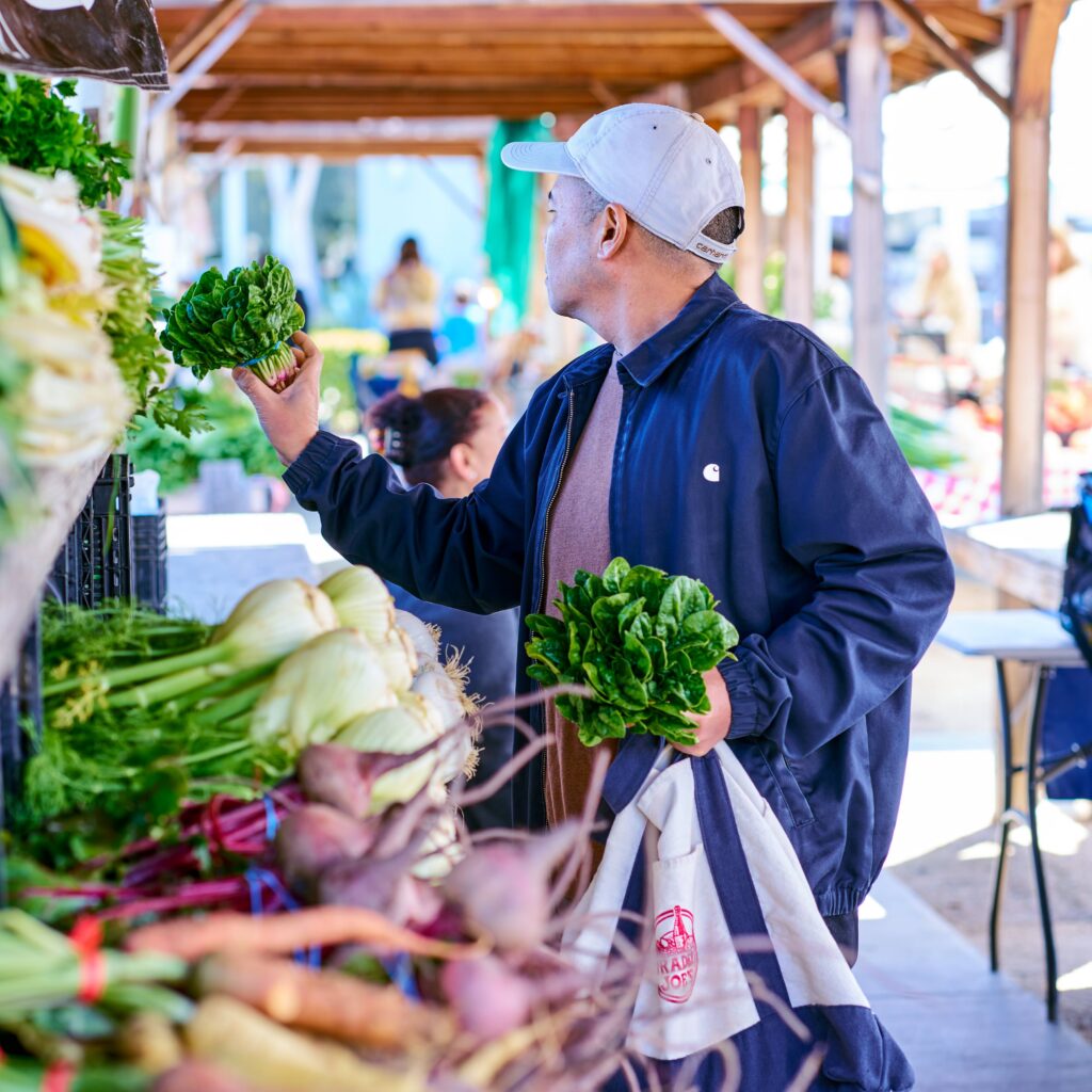 A man wearing a white cap and dark jacket shops at an outdoor market, holding leafy greens in one hand and a reusable bag in the other, surrounded by fresh vegetables.