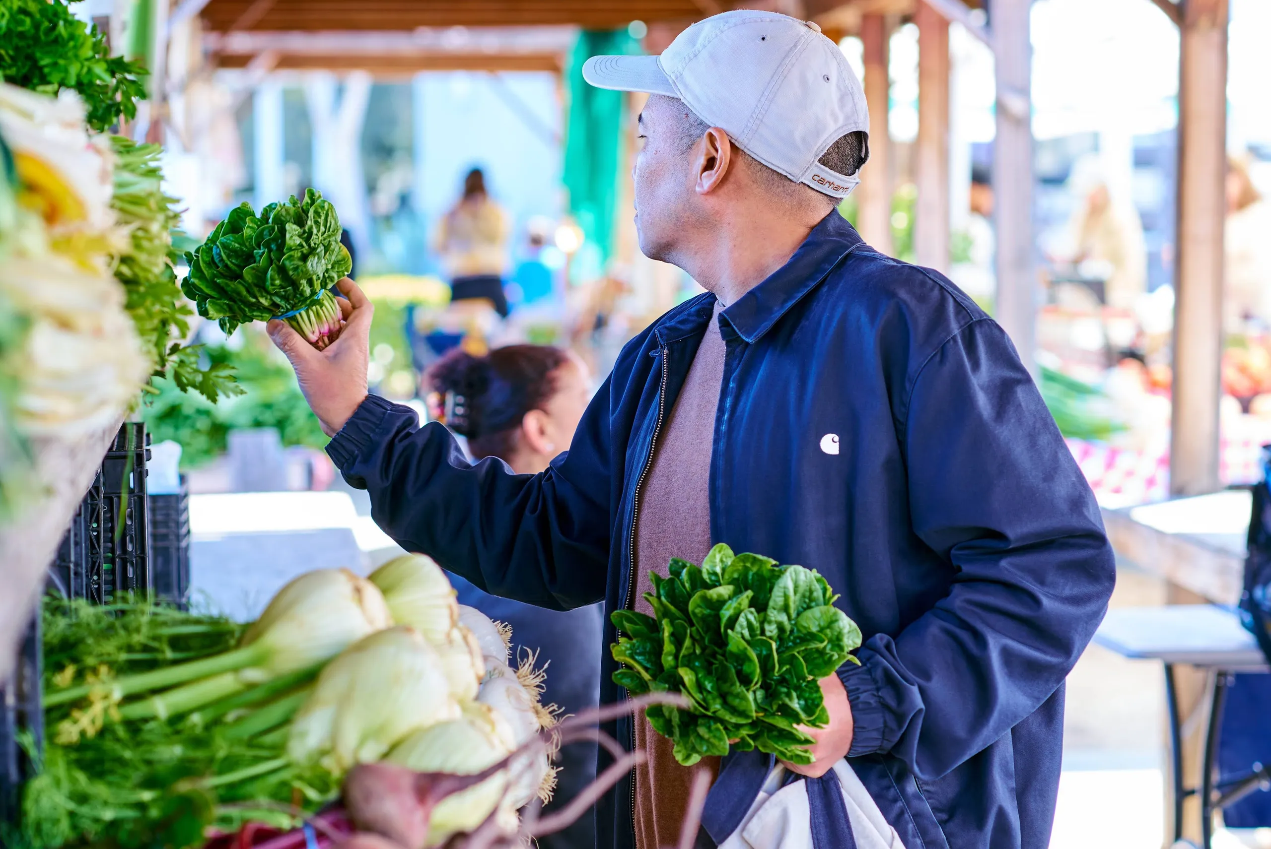 A man wearing a white cap and dark jacket shops at an outdoor market, holding leafy greens in one hand and a reusable bag in the other, surrounded by fresh vegetables.