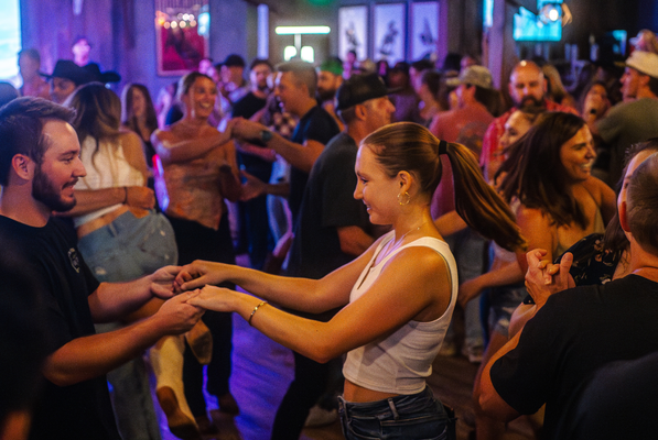 A lively crowd of people dance in pairs at a dimly lit indoor venue. The atmosphere is energetic and joyful, with smiling faces and upbeat body language.