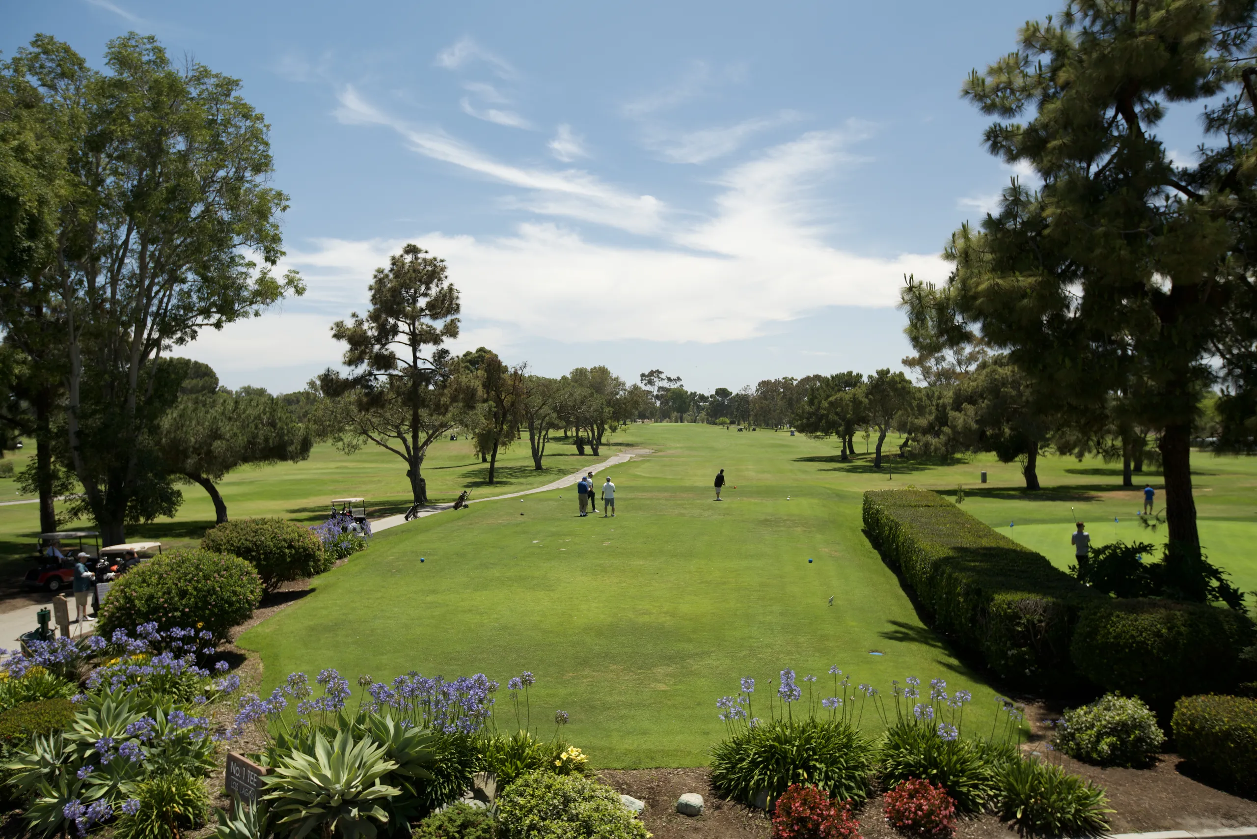 A lush golf course in Mesa Verde, Costa Mesa, with several people playing under a blue sky, surrounded by trees, bushes, and flowers; a manicured lawn stretches into the distance.
