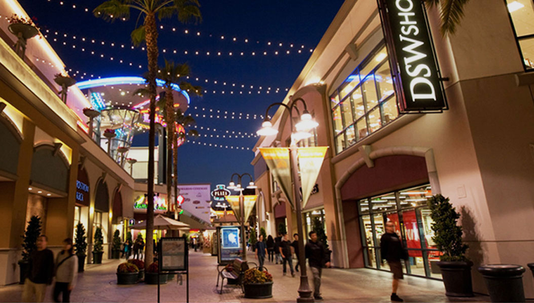 Shoppers walk through a brightly lit outdoor shopping center in the evening, one of the standout Costa Mesa attractions. String lights hang above, and stores like DSW Shoes line both sides of the walkway under a dark blue sky.