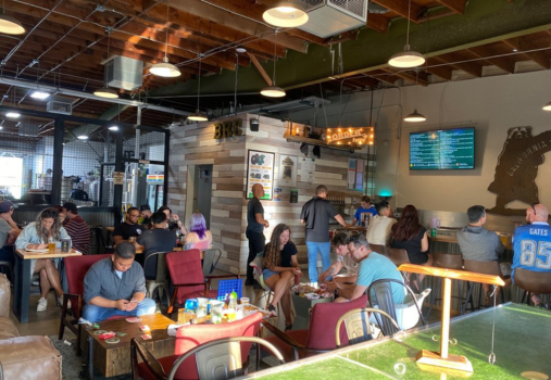 A lively brewery taproom with people sitting at tables, playing games, and socializing—the perfect spot to add to your list of things to do in Costa Mesa. The interior features exposed beams, a wooden bar, a menu board, and a TV screen on the wall.