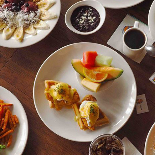 A table set for breakfast at Mesa Verde Costa Mesa features waffles topped with eggs and sauce, a bowl of fruit, sliced bananas with cheese, coffee, sweet potato fries, black beans, and a glass of iced drink.