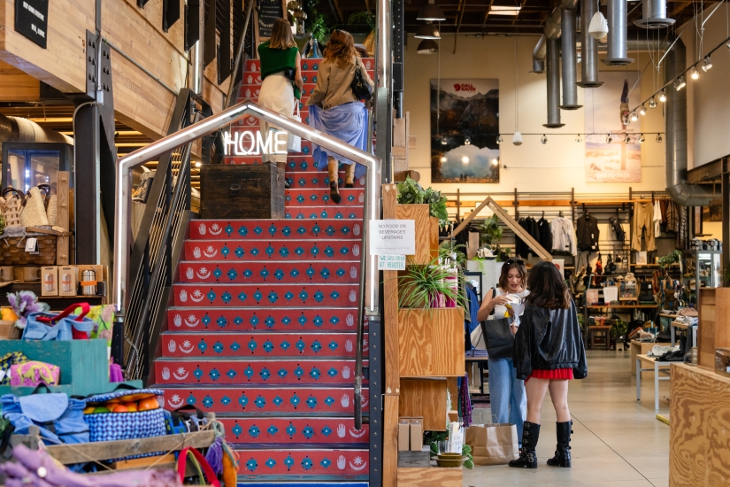 A colorful staircase with a “HOME” sign leads to an upper floor in this bright, modern shopping mall Costa Mesa. Shoppers browse baskets and plants amid wooden displays and striking industrial decor.