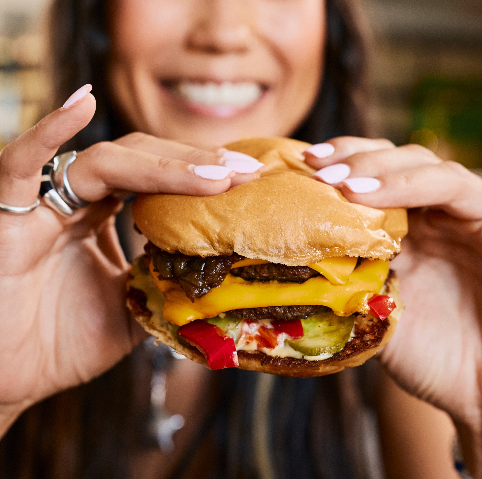A smiling person holding a cheeseburger with pickles, red peppers, melted cheese, and a soft bun, ready to take a bite. Their nails are painted light pink.