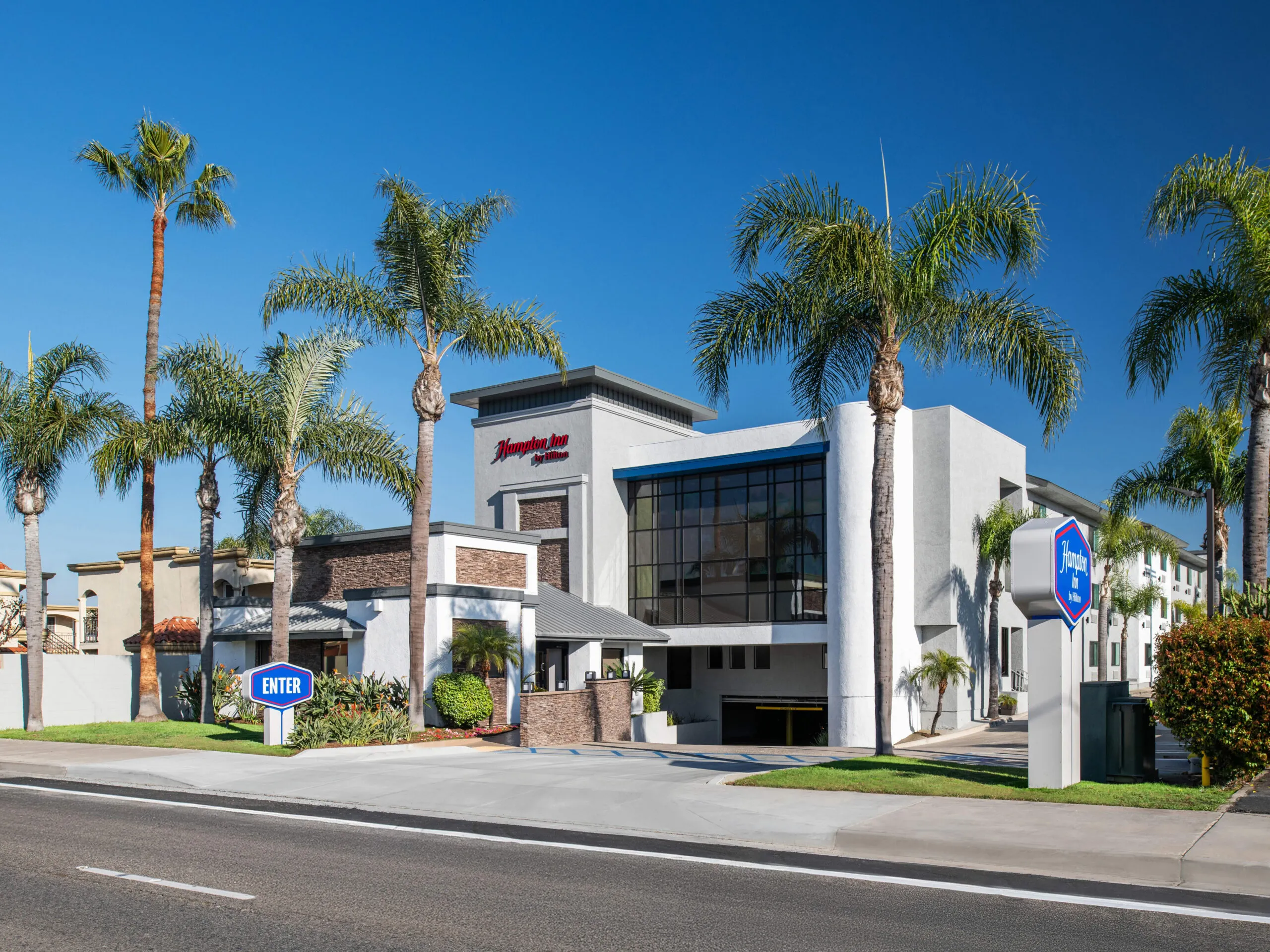 A modern hotel building with large windows and palm trees in front, under a clear blue sky in Mesa Verde, Costa Mesa. A blue "Enter" sign and a "Hampton Inn" sign are visible near the driveway entrance.