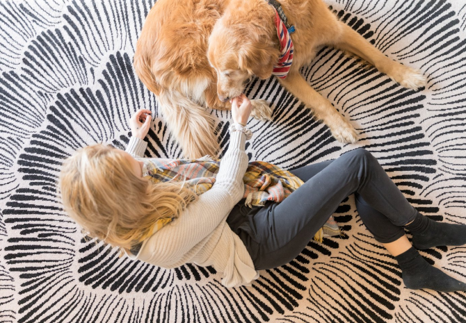 A woman with blonde hair sits on a patterned black-and-white rug, smiling and holding the paw of her golden retriever—one of the friendly faces you might meet while exploring Costa Mesa attractions. The dog wears a colorful bandana.