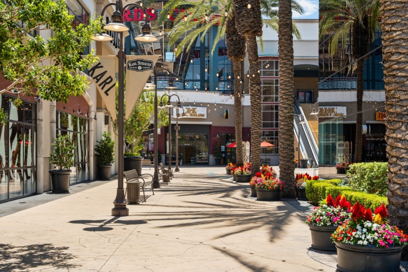 Outdoor shopping plaza in Costa Mesa with palm trees, flower planters, and storefronts—including one with a KARL sign. Strings of lights hang above, casting shadows on the sunny pavement at this vibrant shopping mall.