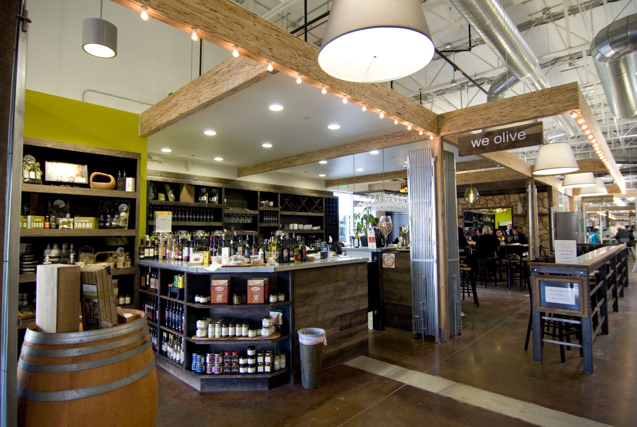 A modern market stall at Mesa Verde Costa Mesa, with shelves of bottles and jars, a wine barrel in front, and a wooden counter. Overhead lights hang from the ceiling as people relax at tables in the background.