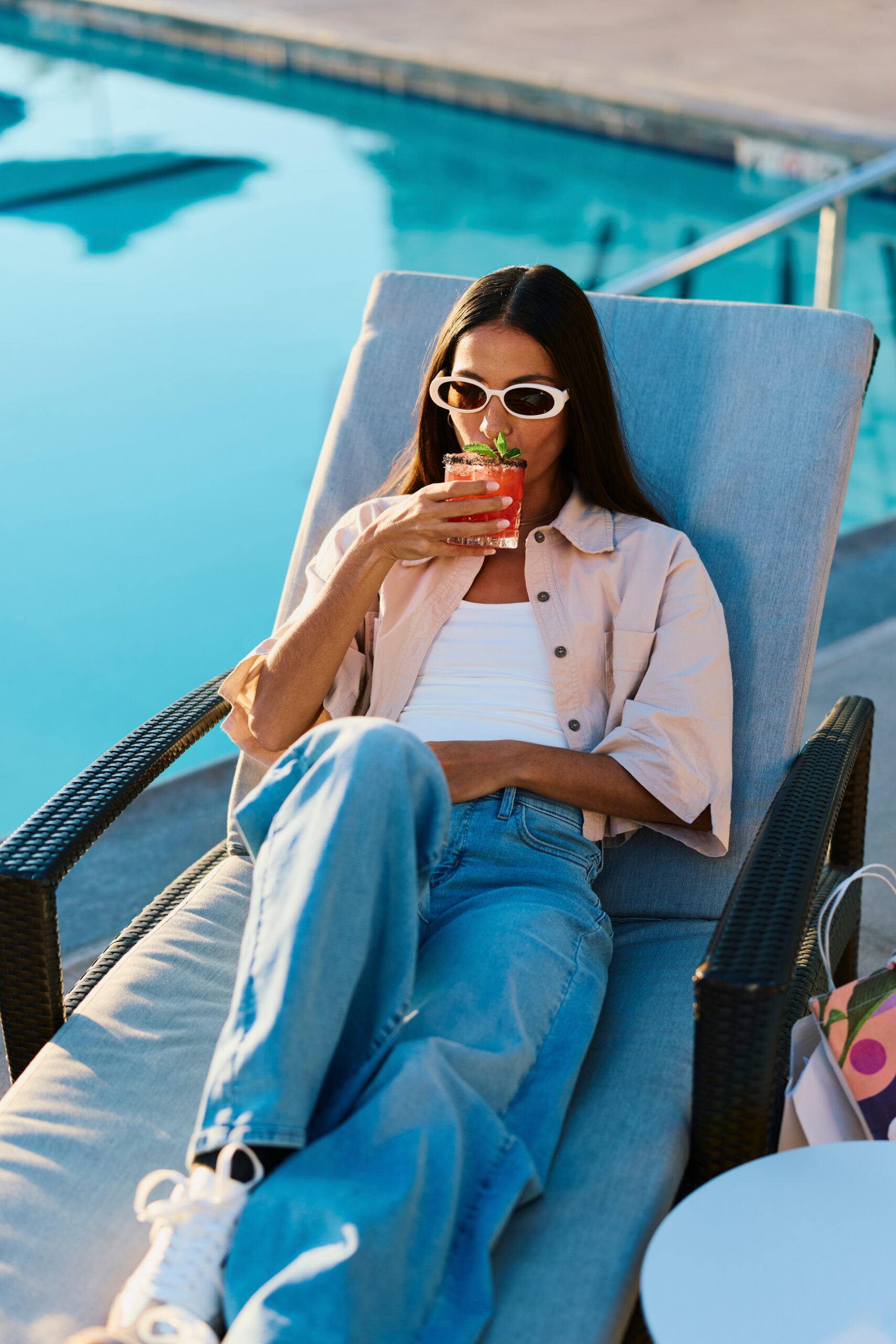 A woman in sunglasses relaxes on a poolside lounge chair, sipping a pink drink garnished with mint. Dressed casually, she enjoys a sunny day by the water, soaking up one of the top Costa Mesa attractions.