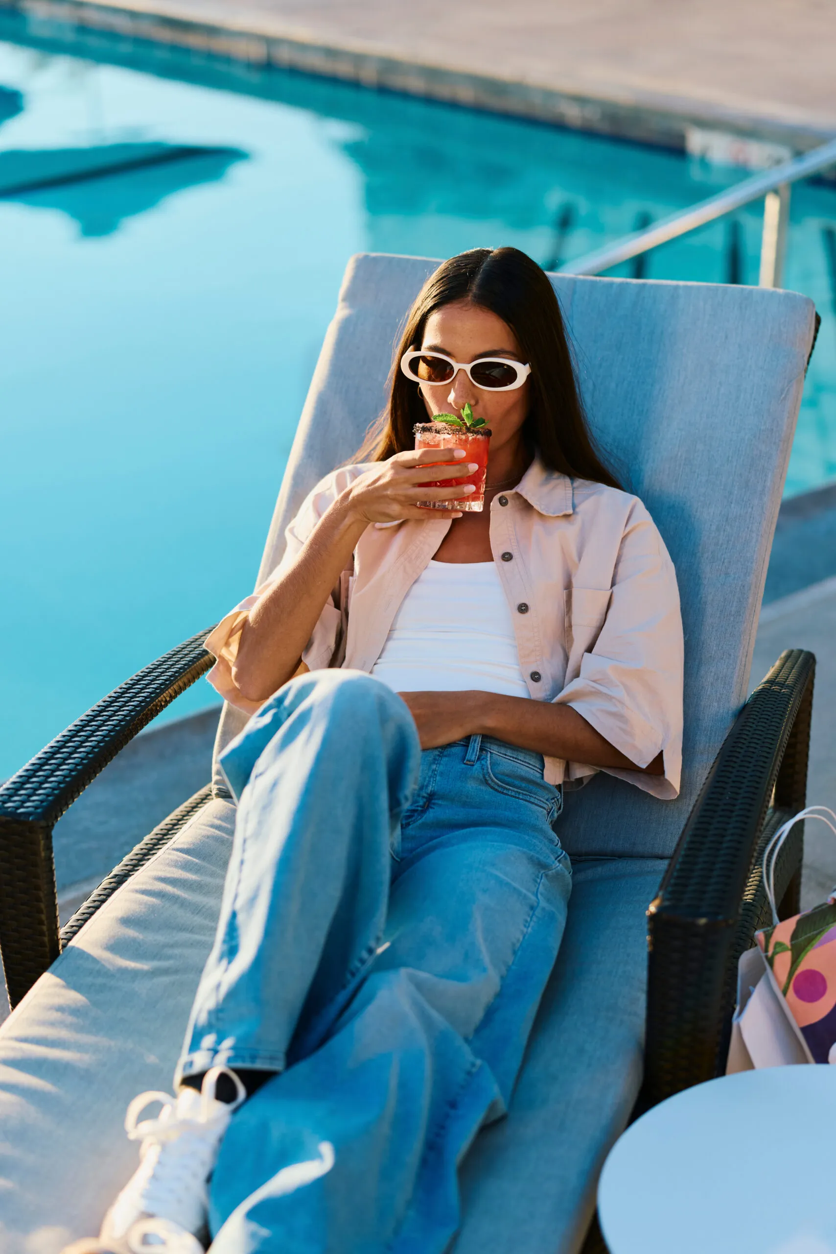 A woman in sunglasses relaxes on a poolside lounge chair, sipping a pink drink garnished with mint. Dressed casually, she enjoys a sunny day by the water, soaking up one of the top Costa Mesa attractions.