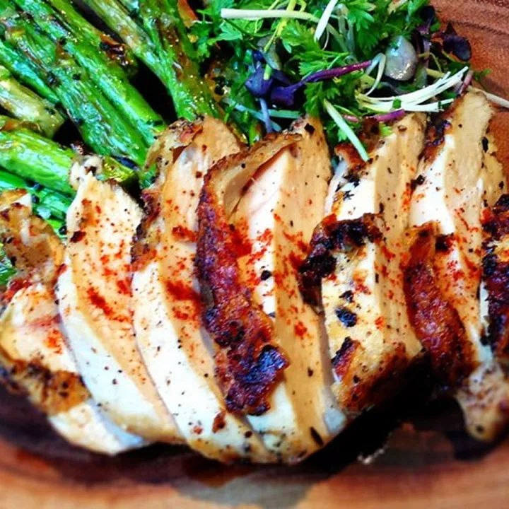Sliced grilled chicken breast seasoned with spices, served with roasted asparagus and a garnish of fresh mixed greens on a wooden plate at Mesa Verde Costa Mesa.