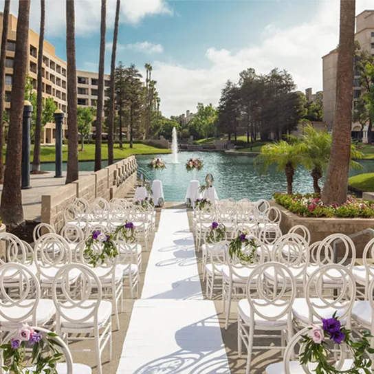 Rows of white chairs with floral decorations are arranged along an aisle facing a small lake and fountain at Avenue of the Arts Costa Mesa, surrounded by trees and buildings under a blue sky, set up for an outdoor wedding ceremony.