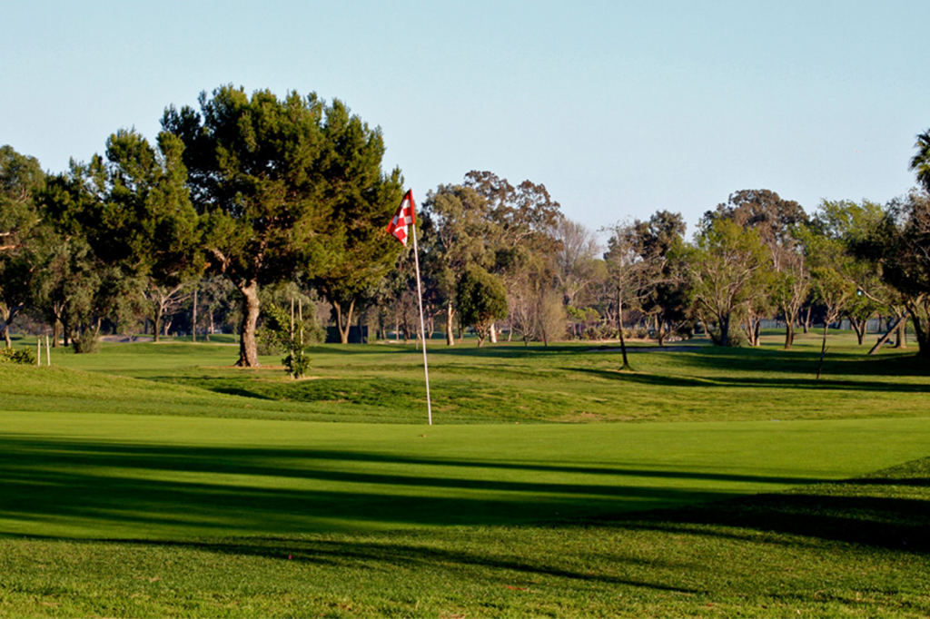 Aerial view of a golf course with green fairways, scattered trees, ponds, a clubhouse with red-tiled roof, golf carts, and a parking lot; cityscape and blue sky in the background.