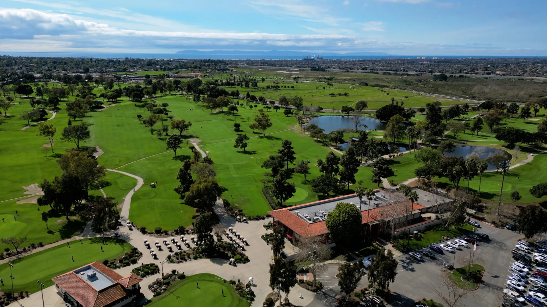 Aerial view of a golf course with green fairways, scattered trees, ponds, a clubhouse with red-tiled roof, golf carts, and a parking lot; cityscape and blue sky in the background.