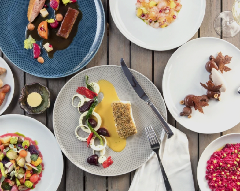 A variety of elegantly plated gourmet dishes on a table in Costa Mesa, including fish with sauce, a colorful salad, desserts, and butter with bread, all arranged artfully on different plates.