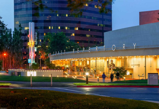A modern theater building labeled REPERTORY is brightly lit at dusk, with people entering. The Segerstrom Performing Arts Center in Costa Mesa stands nearby, with a tall sign, glass high-rise, trees, and a well-kept lawn completing the scene.