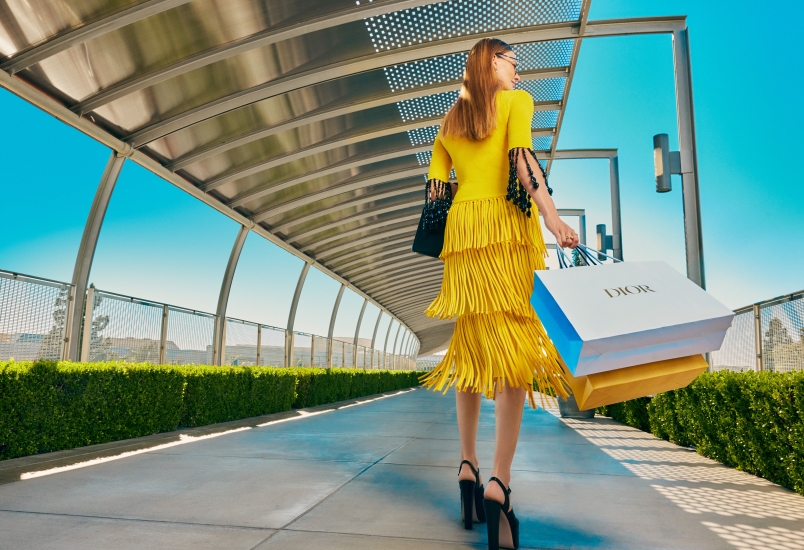 A woman in a bright yellow dress and high heels walks along a modern outdoor walkway at the shopping mall Costa Mesa, carrying several shopping bags, including a large white Dior bag, under a blue sky.