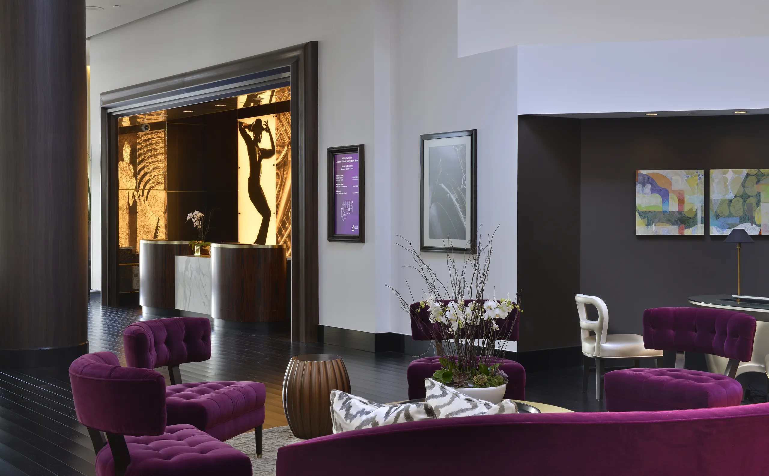 Modern hotel lobby with purple velvet chairs, a white orchid centerpiece, abstract artwork on walls, and a lit reception desk featuring a golden art piece behind it. Located near Avenue of the Arts Costa Mesa, the space has a sleek and contemporary design.