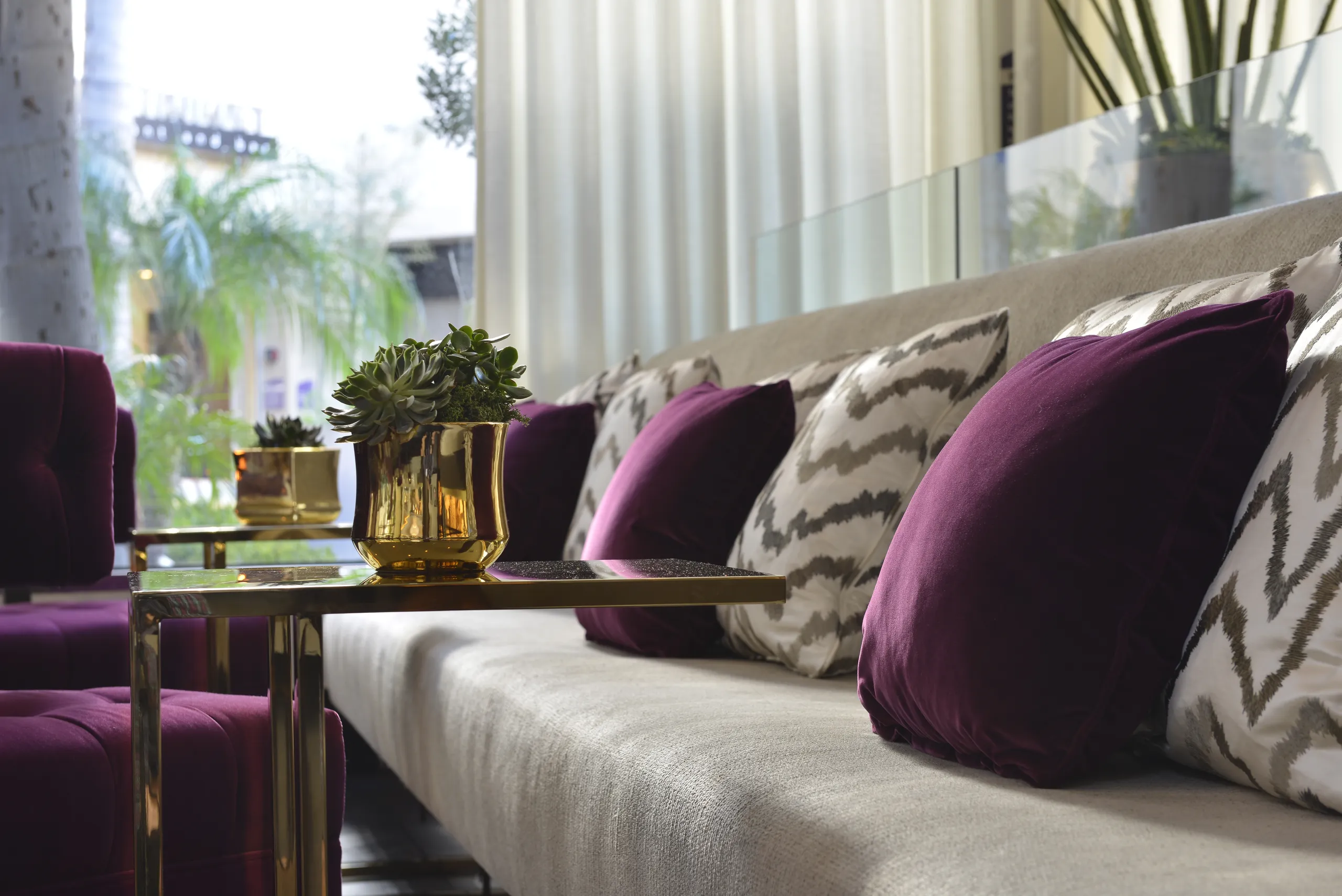 Modern living room with a beige sofa, purple and patterned pillows, a gold side table, and a small potted plant. Light streams in through sheer curtains, and greenery is visible outside the window—capturing the sophisticated ambiance often found near Avenue of the Arts Costa Mesa.