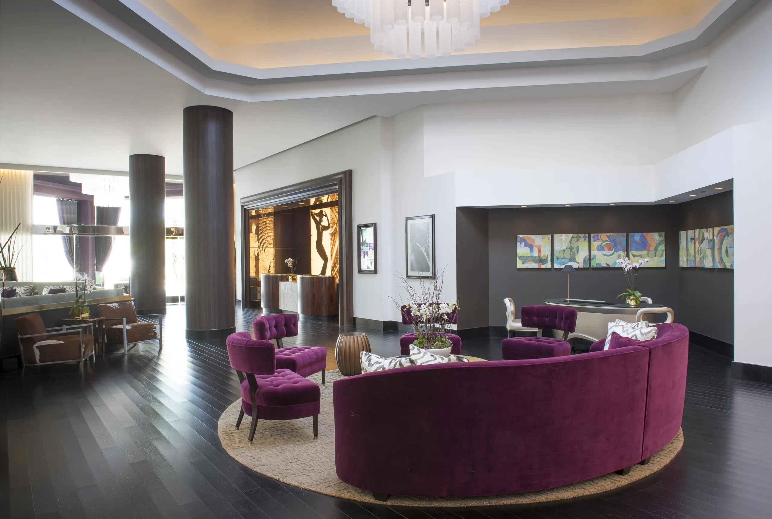 Modern hotel lobby with a curved purple velvet sofa, matching chairs, round rug, dark wood floors, and contemporary art on the walls. Large windows and architectural columns enhance the spacious, stylish atmosphere—an inviting retreat in the heart of Avenue of the Arts Costa Mesa.