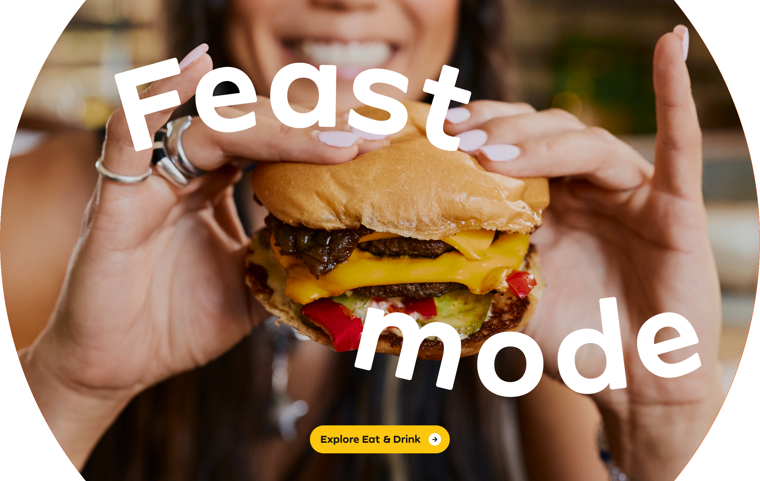 Close-up of a person holding a cheeseburger with lettuce, tomato, and onions in Costa Mesa. The words Feast mode are overlaid on the image, and a yellow button reads Explore Eat & Drink.