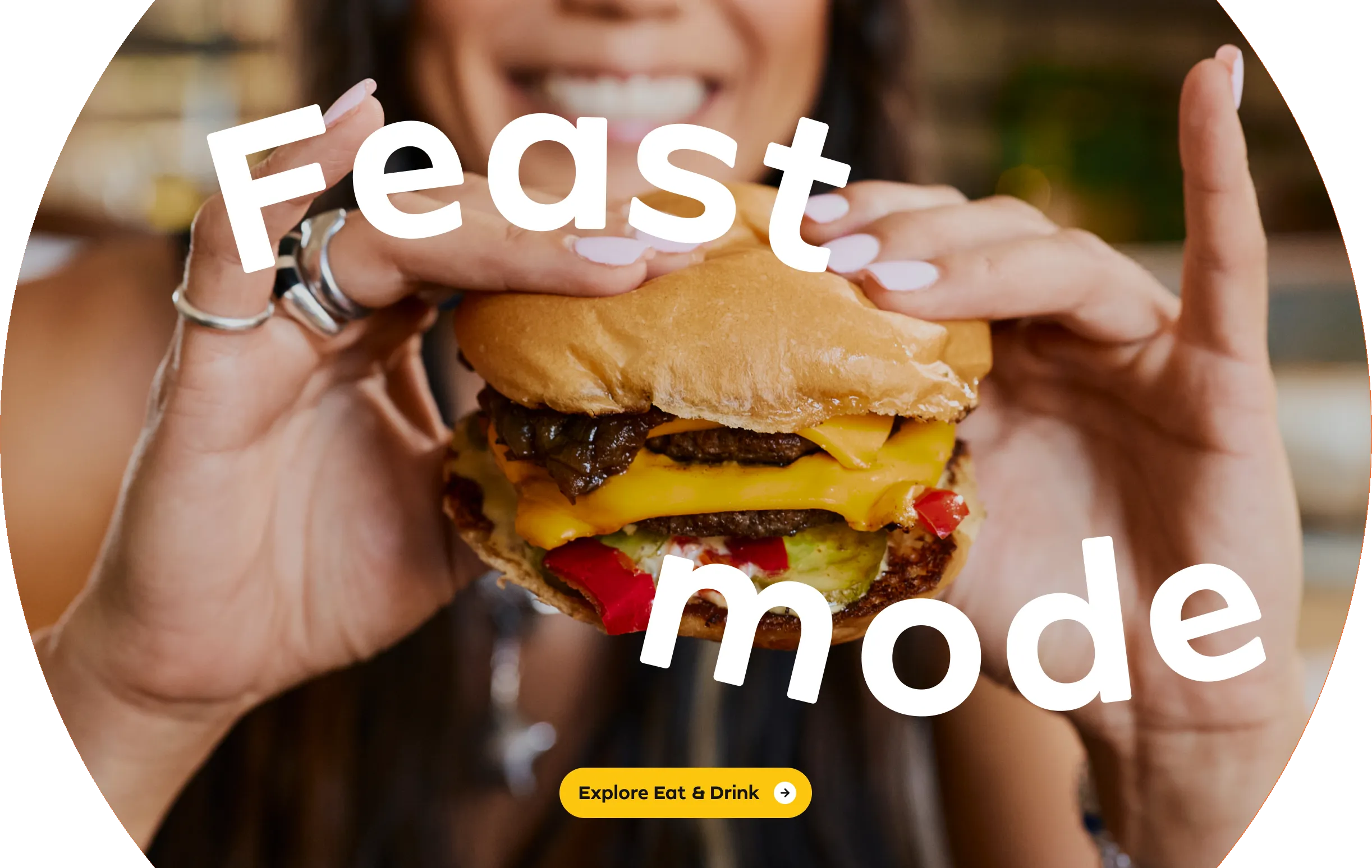 Close-up of a person holding a cheeseburger with lettuce, tomato, and onions in Costa Mesa. The words Feast mode are overlaid on the image, and a yellow button reads Explore Eat & Drink.