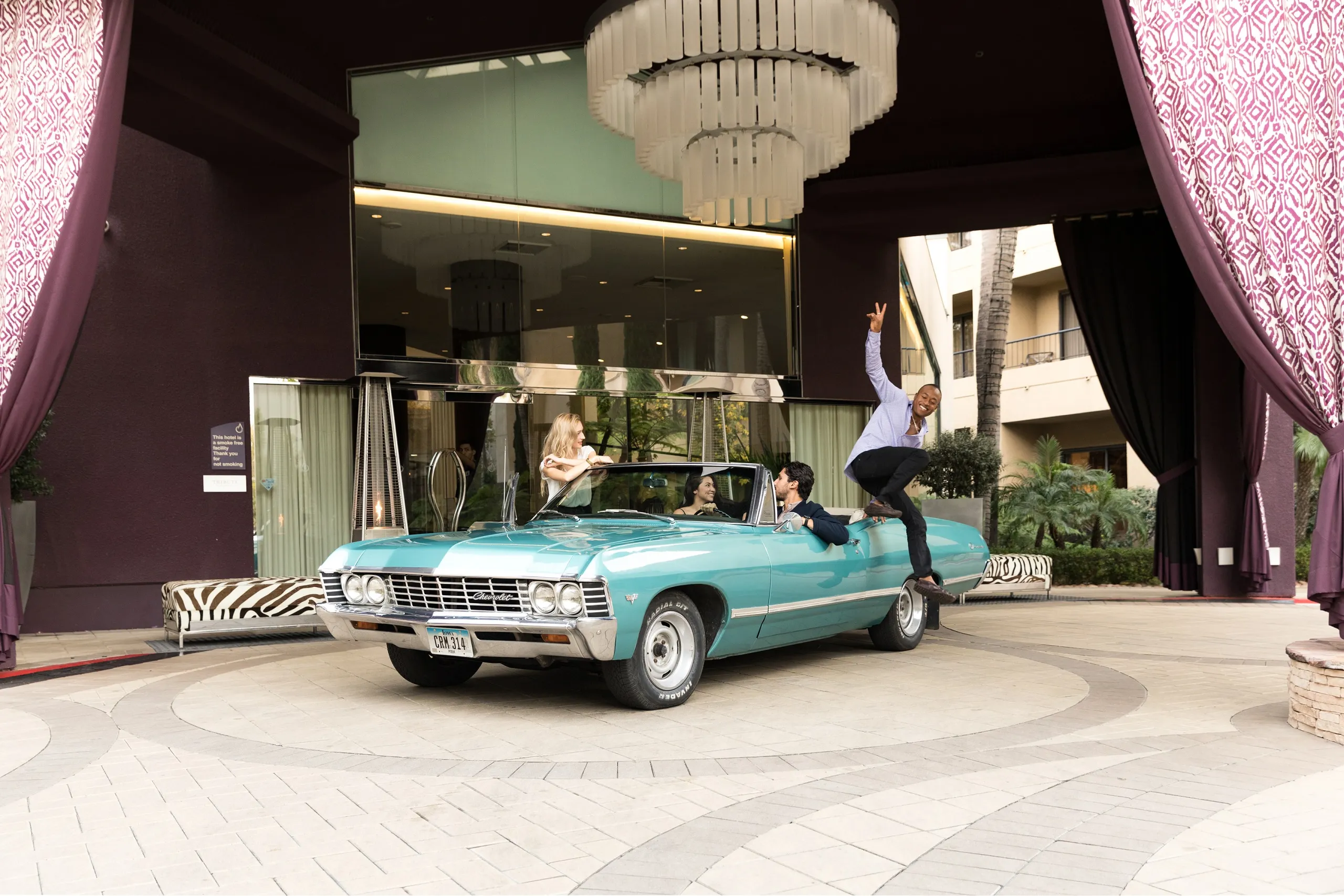 Four people enjoy a classic turquoise convertible parked under a grand entrance near Avenue of the Arts Costa Mesa, with large curtains and chandeliers. One person stands beside the car, raising their hand in excitement.