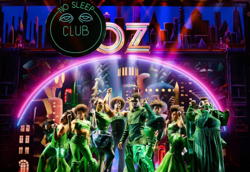 Performers in vibrant green costumes dance energetically on a colorful stage with OZ in bold yellow letters behind them, bringing South Coast Plaza shopping excitement to life. A neon “No Sleep Club” sign is overlaid in the upper left corner.