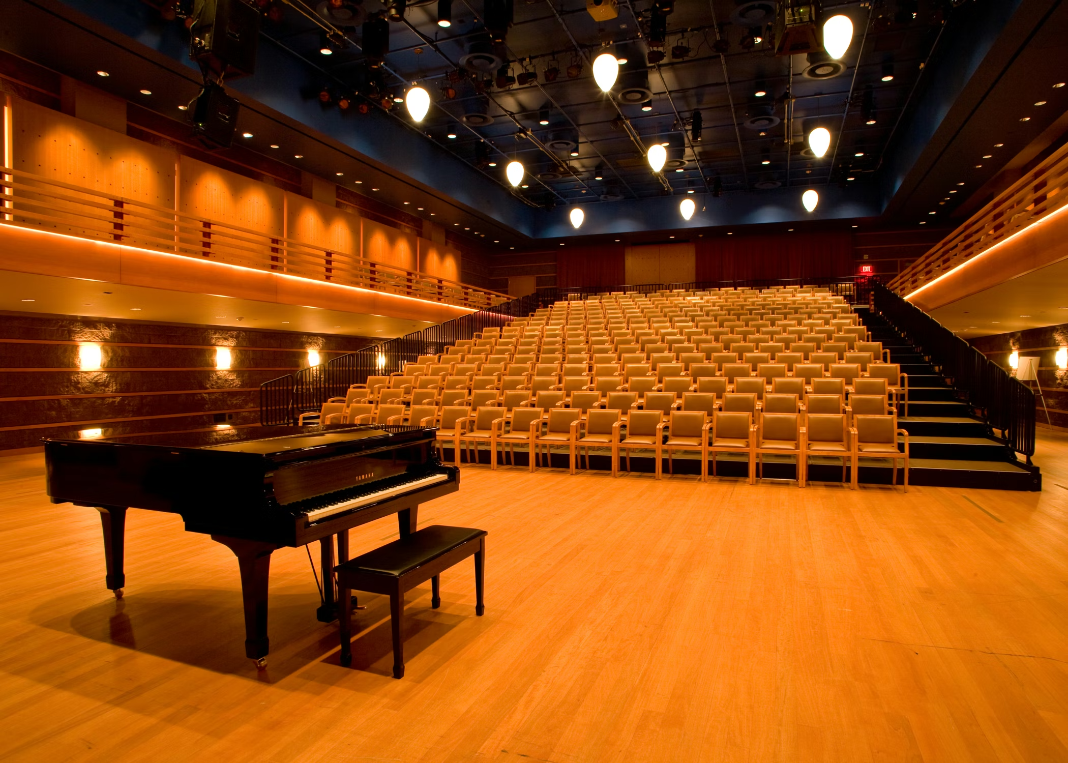 A grand piano with a bench sits on a wooden stage in the empty, well-lit Samueli Theater Costa Mesa auditorium, surrounded by rows of beige seats and modern ceiling lights.