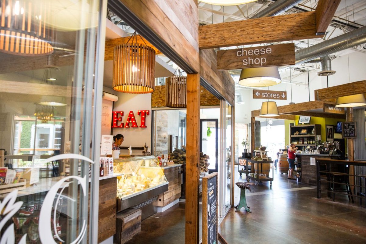 A rustic cheese shop interior with wooden beams, a glass display case of cheeses, and a sign that says cheese shop. People are seen in the background near tables and a counter in the cozy Mesa Verde Costa Mesa market setting.