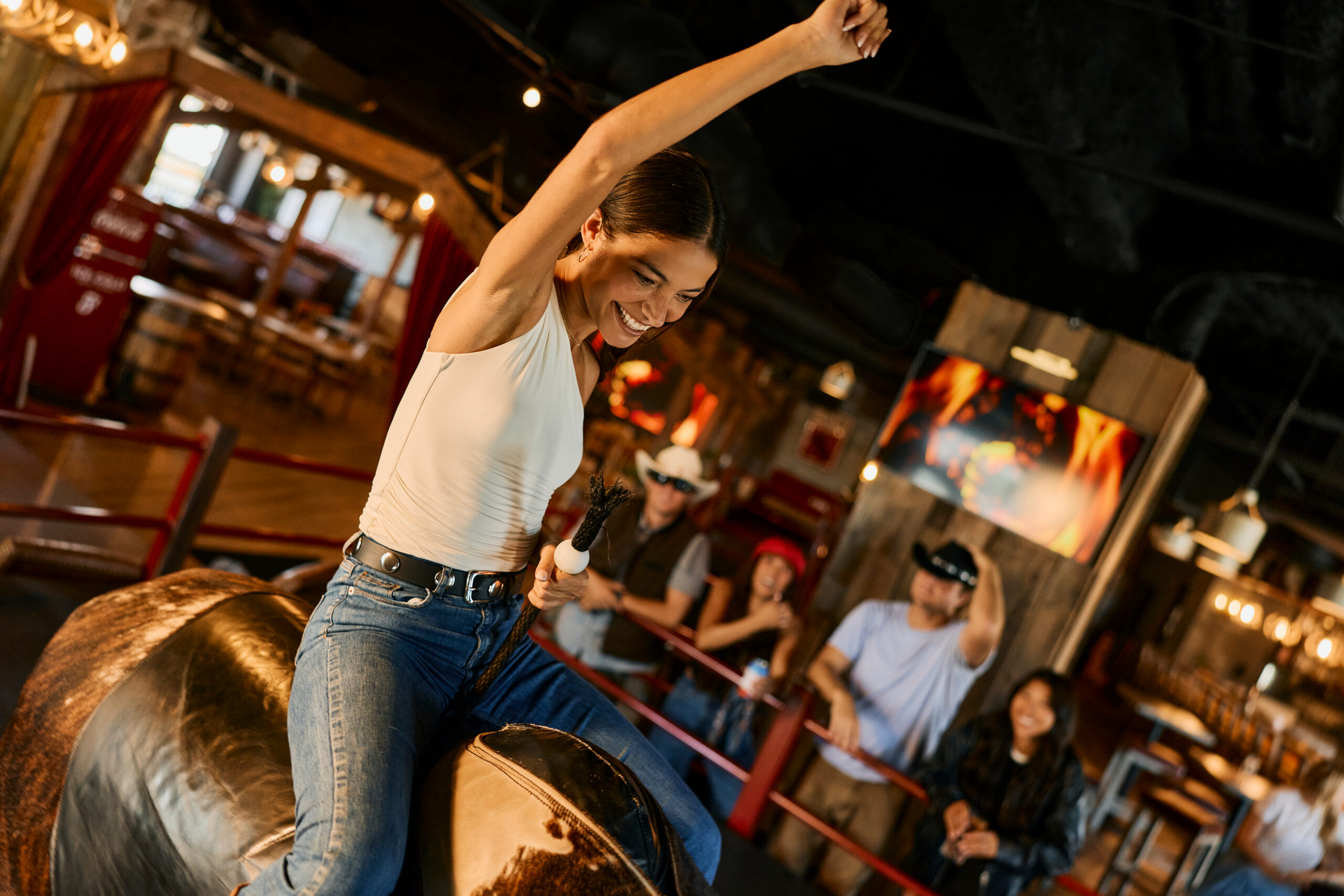 A smiling woman in a white tank top and jeans rides a mechanical bull indoors, raising one arm triumphantly, while a group of people in the background watch and cheer—one of the exciting things to do in Costa Mesa.