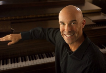 A smiling bald man in a dark shirt sits at a piano with one arm resting on the keys, looking at the camera in a warmly lit room after a day of south coast plaza shopping.
