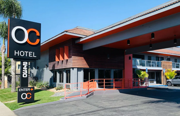 Modern hotel exterior in Mesa Verde Costa Mesa with a large black and orange OC Hotel sign, orange railings, wood accents, expansive windows, and a covered entrance. The address 2430 is clearly visible on a sign.