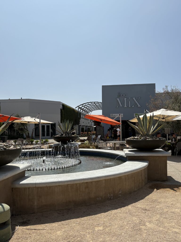 A round fountain with water jets is in the foreground, surrounded by large agave plants. Behind it is a modern building at The OC Mix in Mesa Verde Costa Mesa, featuring outdoor seating and orange umbrellas under a clear sky.