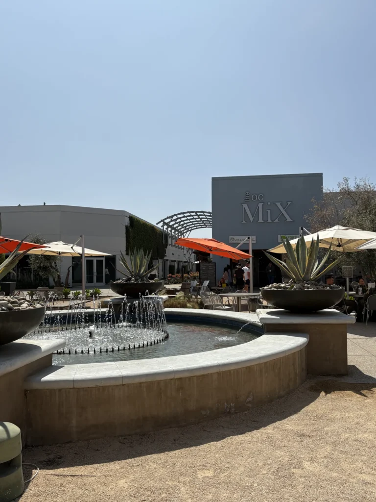 A round fountain with water jets is in the foreground, surrounded by large agave plants. Behind it is a modern building at The OC Mix in Mesa Verde Costa Mesa, featuring outdoor seating and orange umbrellas under a clear sky.