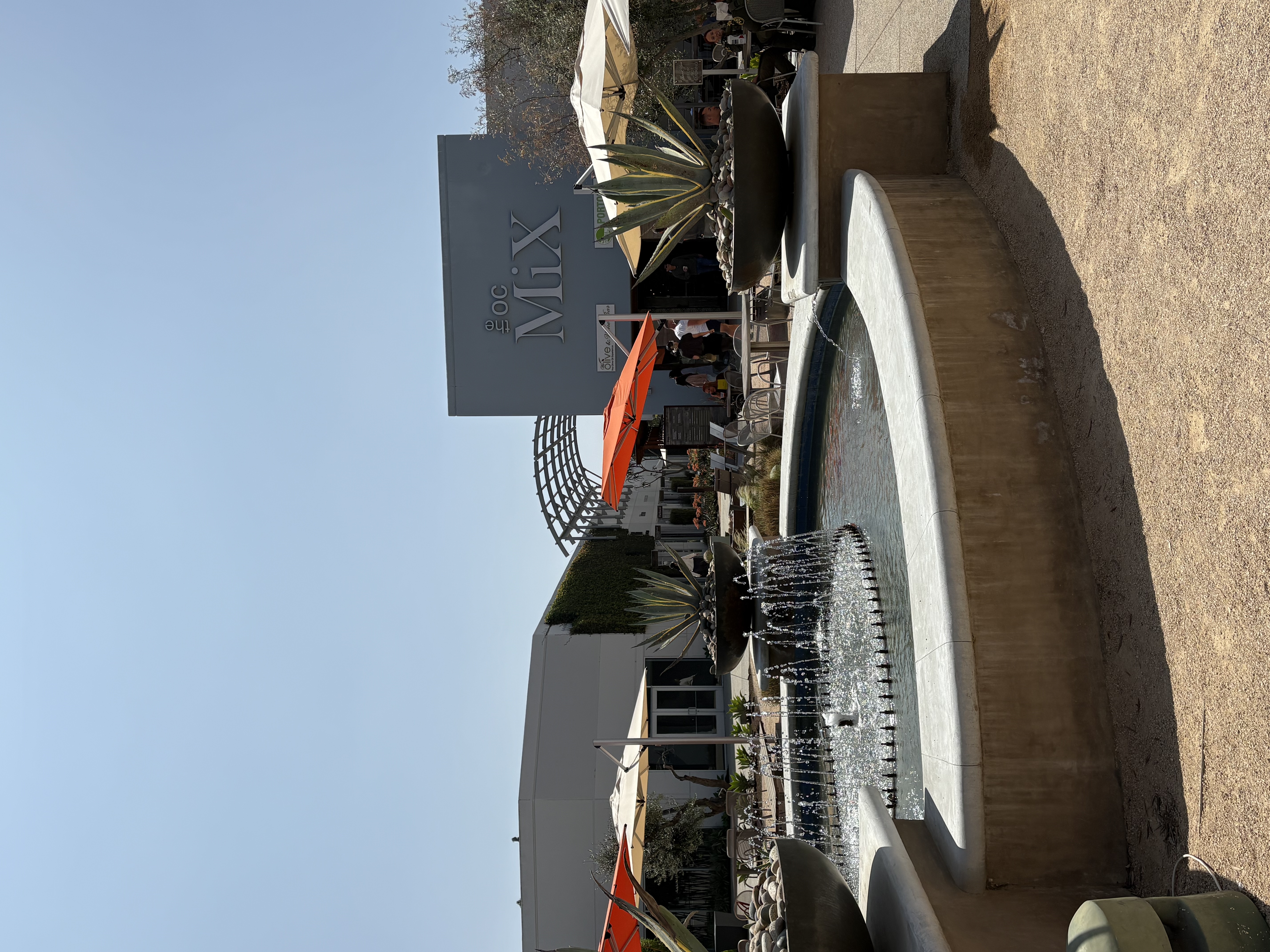 A round fountain with water jets is in the foreground, surrounded by large agave plants. Behind it is a modern building at The OC Mix in Mesa Verde Costa Mesa, featuring outdoor seating and orange umbrellas under a clear sky.