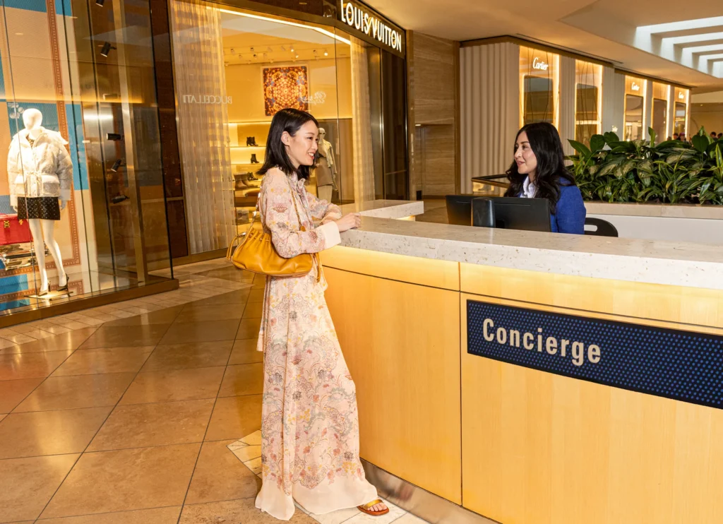 A woman in a long floral dress stands at a concierge desk in South Coast Plaza shopping mall, speaking to a receptionist. Luxury stores including Louis Vuitton and Cartier are visible in the background.