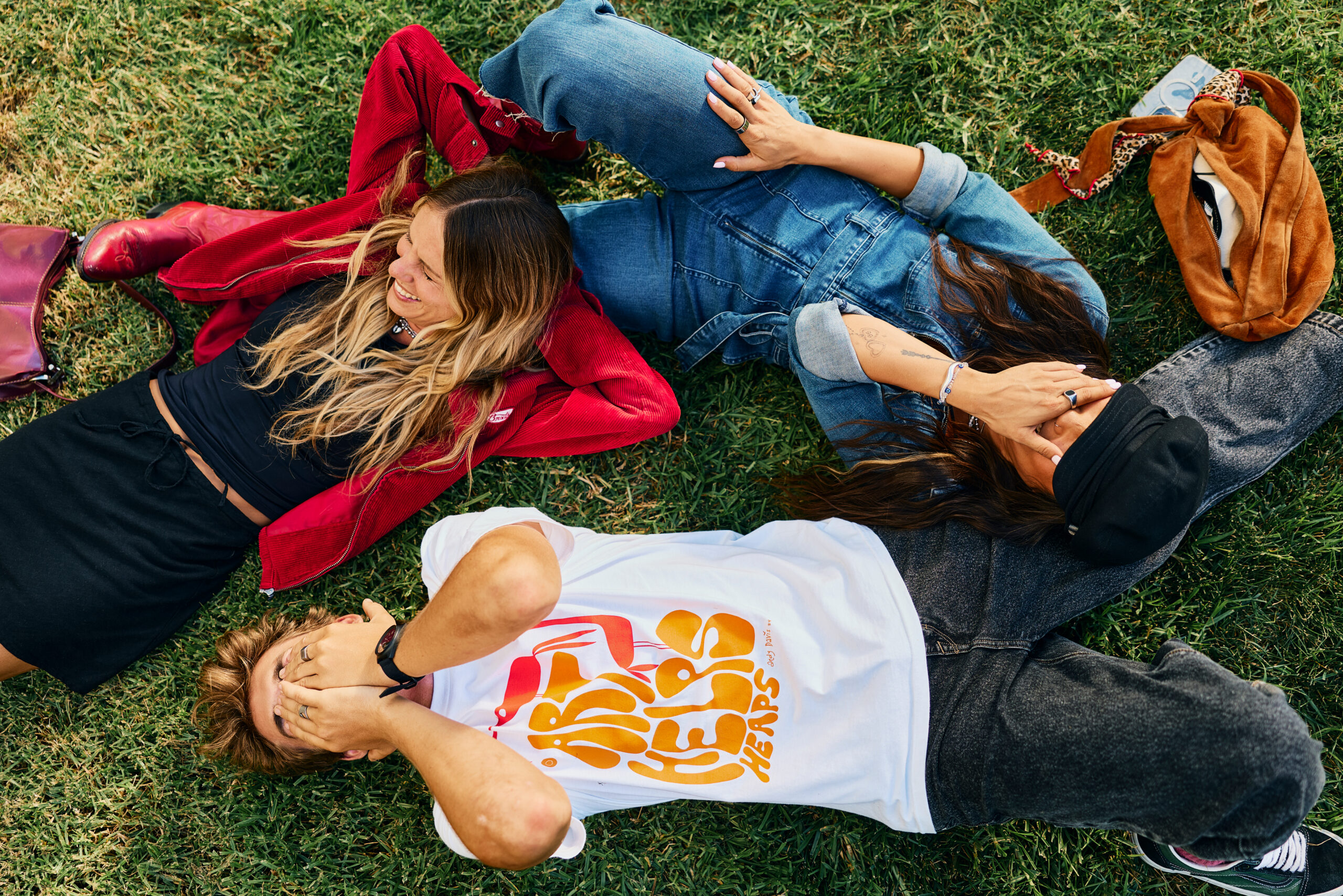 Three people lie on grass in Costa Mesa, laughing and playfully covering their faces. One wears a red jacket, another denim, and the third a white graphic t-shirt. A brown bag and red purse rest nearby at Mesa Verde Park.