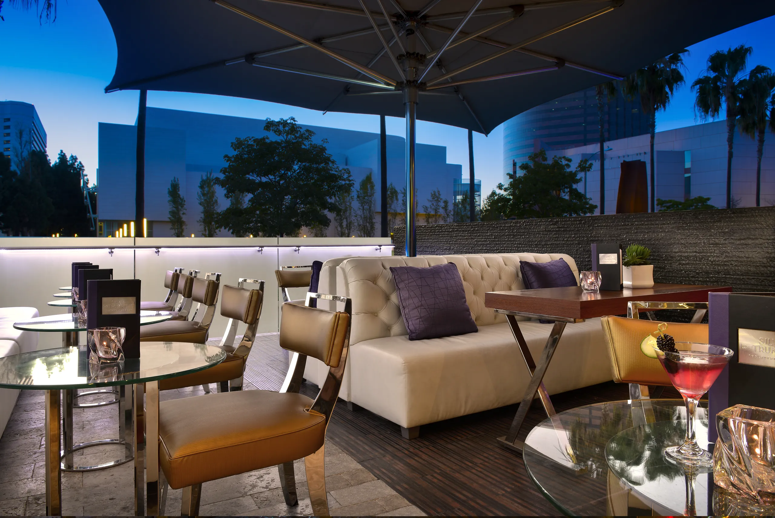 Outdoor lounge with modern chairs, glass tables, a white sofa, drinks on tables, and city buildings in the background at dusk—perfectly capturing the vibrant atmosphere near Avenue of the Arts Costa Mesa. A large umbrella provides shade above the seating area, making it an inviting spot to relax and enjoy the evening.